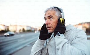 Mature male runner with gloves putting on headphones outdoors on the bridge in Prague city.