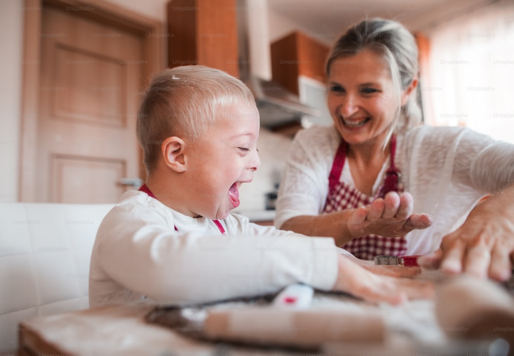 Ein lachendes behindertes Down-Syndrom-Kind und seine Mutter mit karierten Schürzen backen drinnen in einer Küche und haben Spaß.