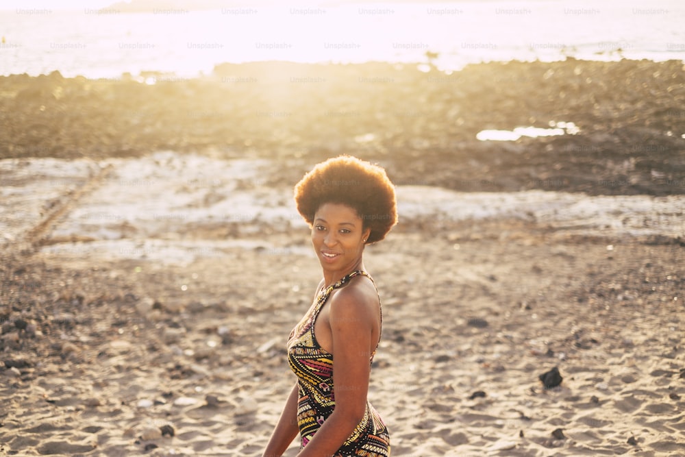 Beautiful afro hair skin race lady on the beach with sunset and backlight in background enjoying outdoor leisure activity. Traditional dress afro style and colors. Beautiful black model