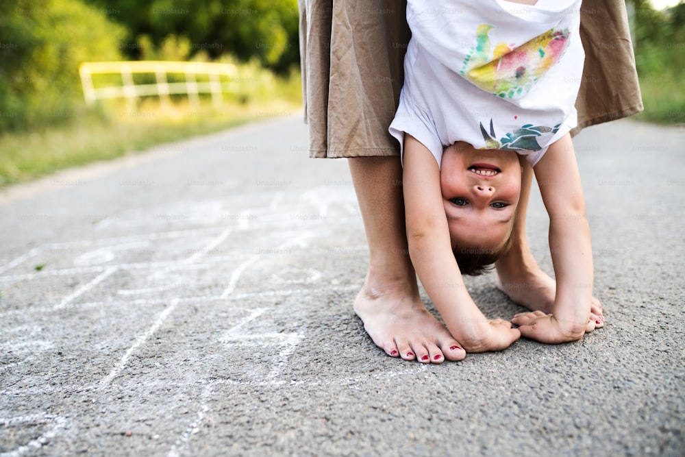 Unrecognizable barefoot mother holding a small son upside down on a road in park on a summer day.