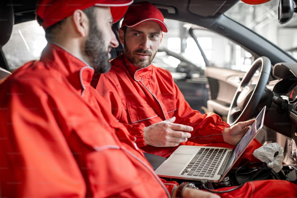 Two male auto mechanics in red uniform diagnosing car with computer sitting on the seats indoors at the car service