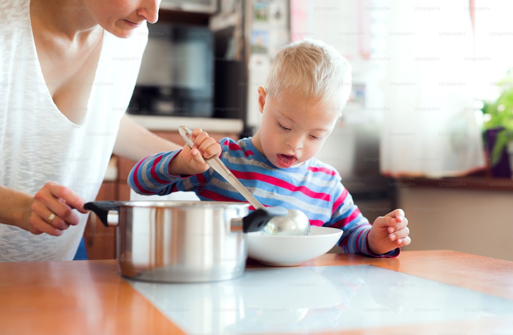 Um menino feliz deficiente com síndrome de down com mãe irreconhecível derramando sopa em um prato dentro de casa, hora do almoço.
