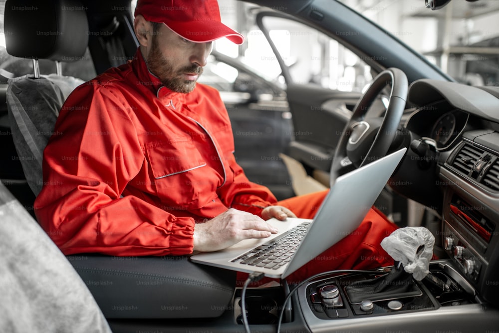 Auto mechanic in red uniform diagnosing car with computer sitting on the driver seat at the car service