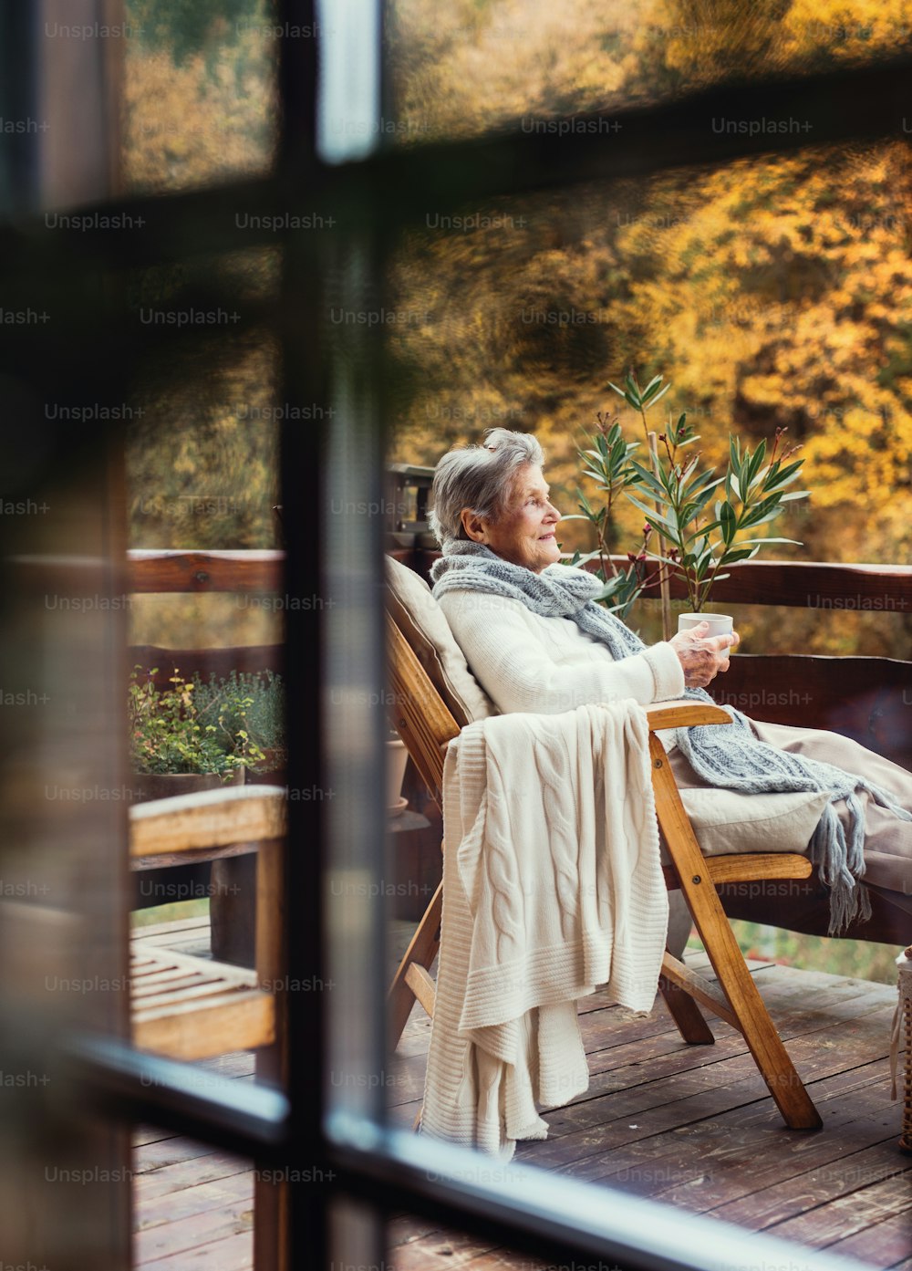秋の晴れた日、屋外のテラスに座ってコーヒーを飲む年配の年配の女性。ガラス越しに撮影。