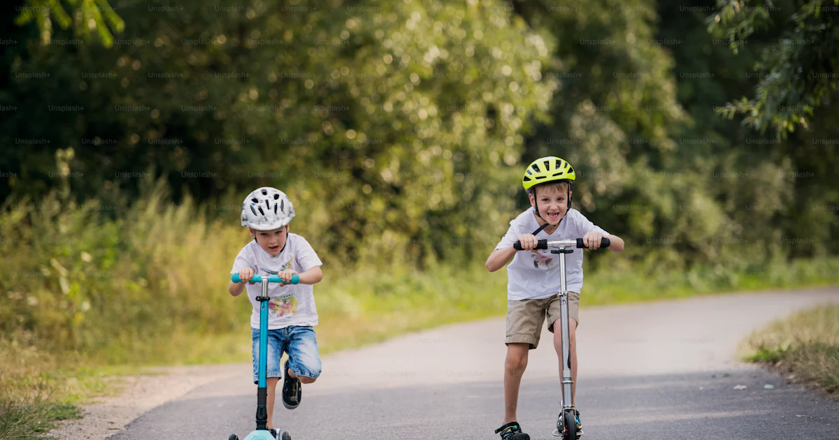 Kids Using Scooters Two cheerful small boys with a helmet riding scooters on a road in park ...