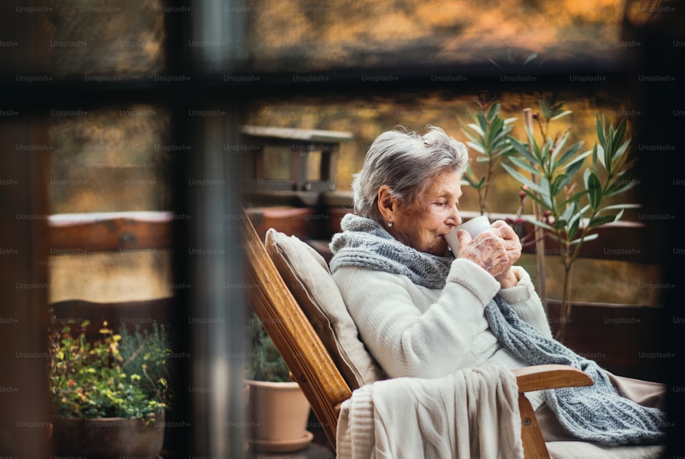 秋の晴れた日、屋外のテラスに座ってコーヒーを飲む年配の年配の女性。ガラス越しに撮影。