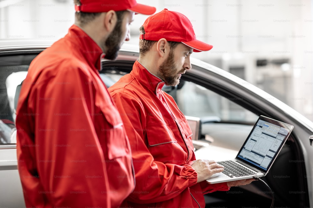 Two male auto mechanics in red uniform diagnosing car with computer standing near the luxury car at the workshop