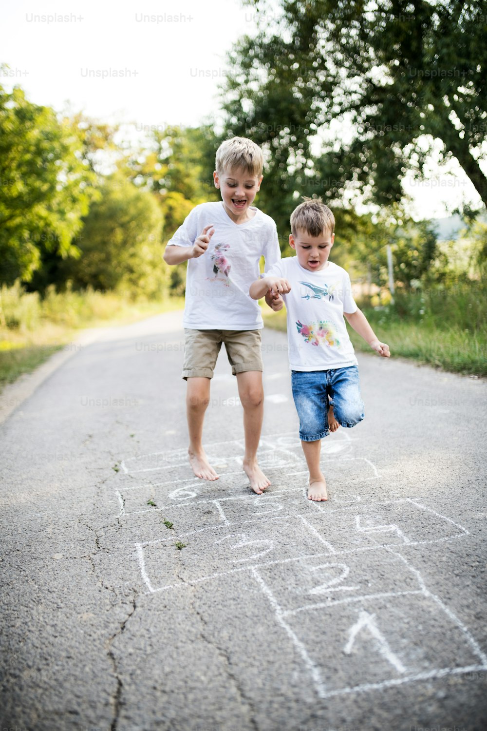 Zwei barfüßige, fröhliche kleine Jungen, die an einem Sommertag auf einer Straße im Park hüpfen.