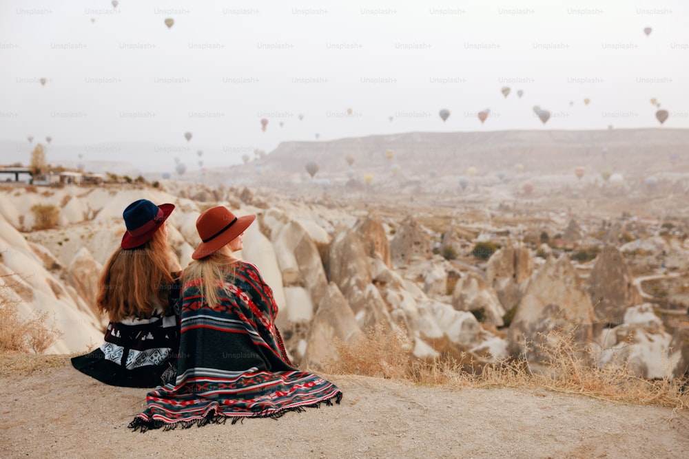 人は旅をします。帽子をかぶった女性が丘の上に座り、トルコのカッパドキアで熱気球を飛ばす様子を楽しんでいます。高解像度