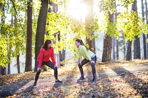 Zwei aktive Läuferinnen strecken sich draußen im Wald in der Herbstnatur nach dem Laufen.