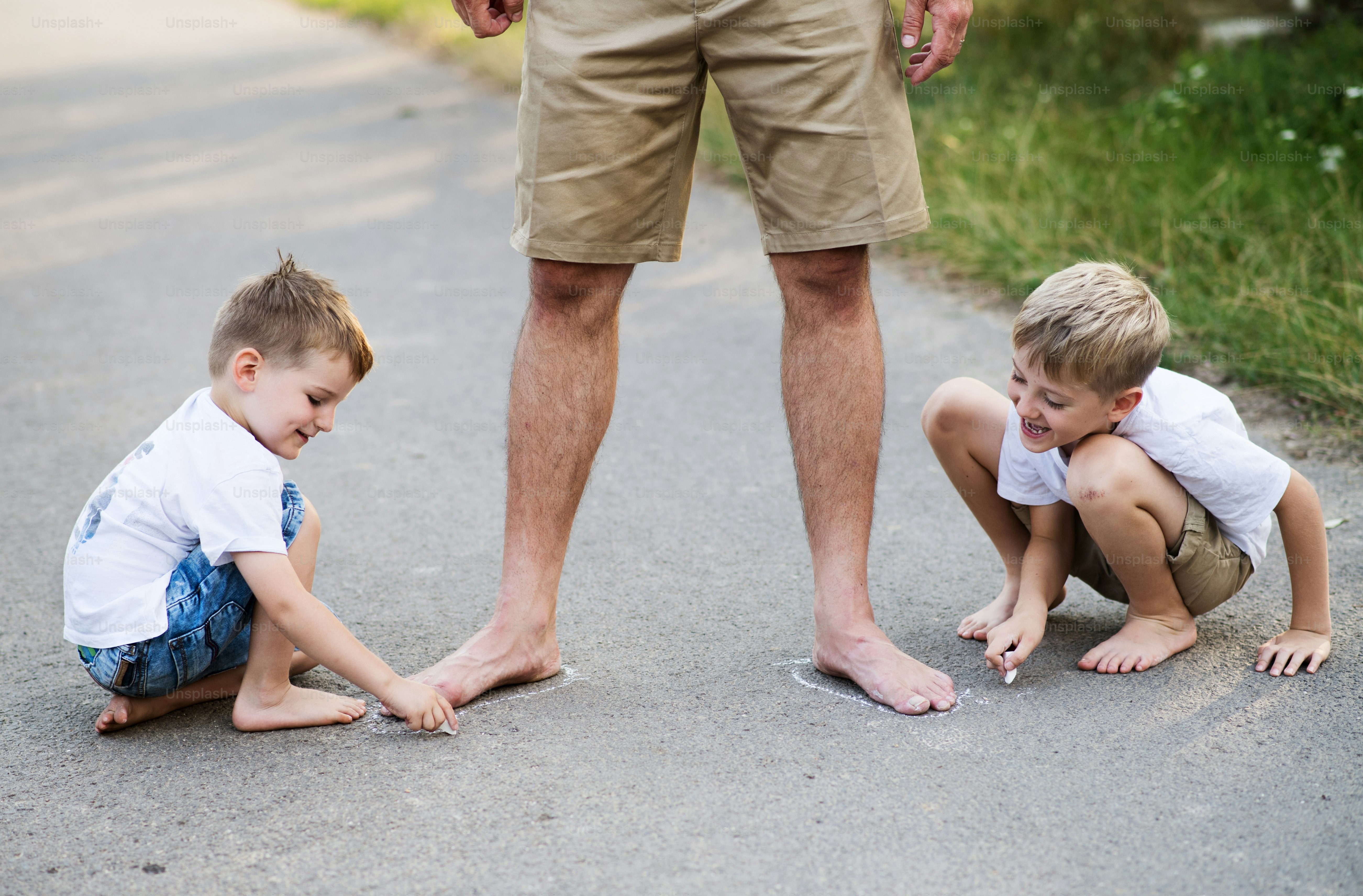 Two small sons playing with unrecognizable father on a road in park on a summer day, drawing with chalk