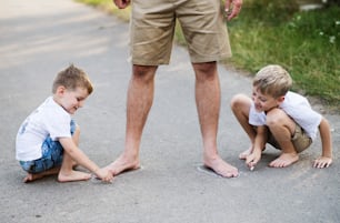 Two small sons playing with unrecognizable father on a road in park on a summer day, drawing with chalk.