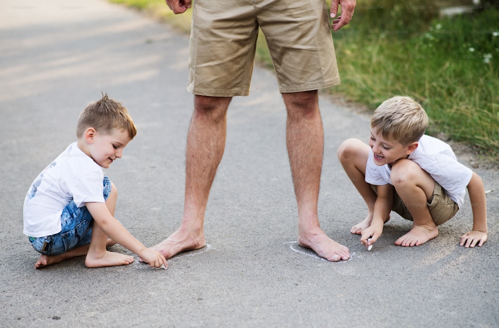 Two small sons playing with unrecognizable father on a road in park on a summer day, drawing with chalk.