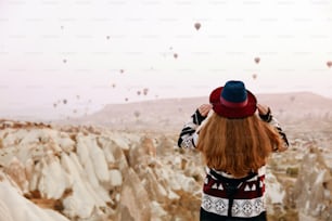 旅行。丘から飛ぶ熱気球を見ている帽子をかぶった美しい女性。カッパドキアの民族衣装を着た女性。高解像度