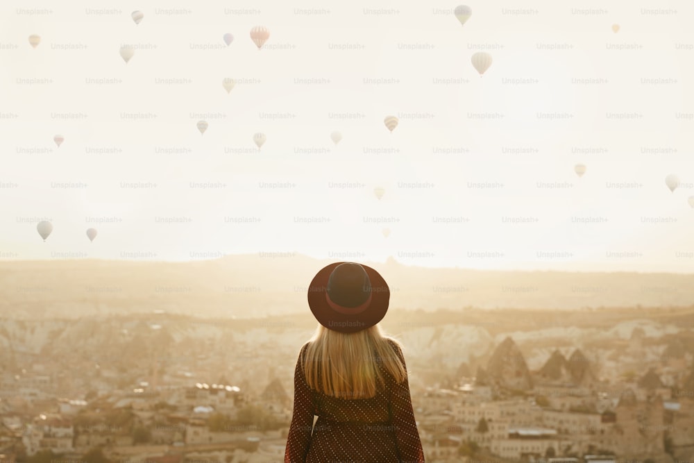 Viajar. Hermosa mujer en sombrero en la colina con globos aerostáticos voladores en el cielo. Mujer viajando a Capadocia. Alta resolución