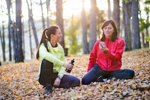 Dos corredoras con teléfono inteligente y botella de agua sentadas en el suelo al aire libre en el bosque en la naturaleza otoñal, descansando.