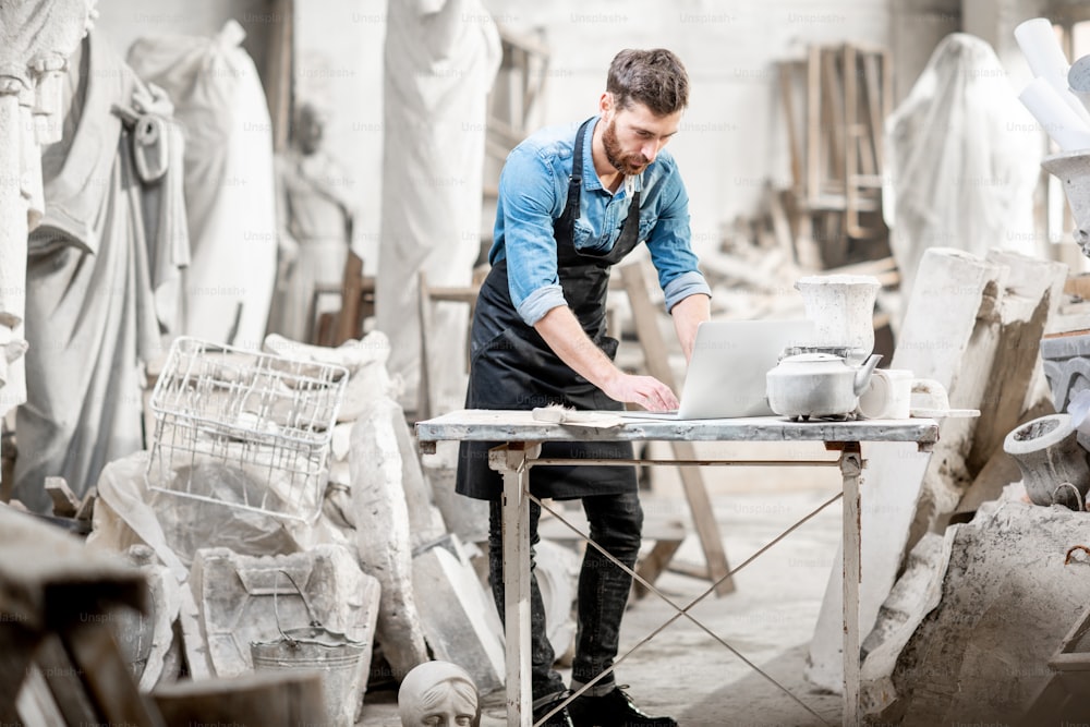 Handsome sculptor working with laptop at the working place in the old studio with sculptures on the background