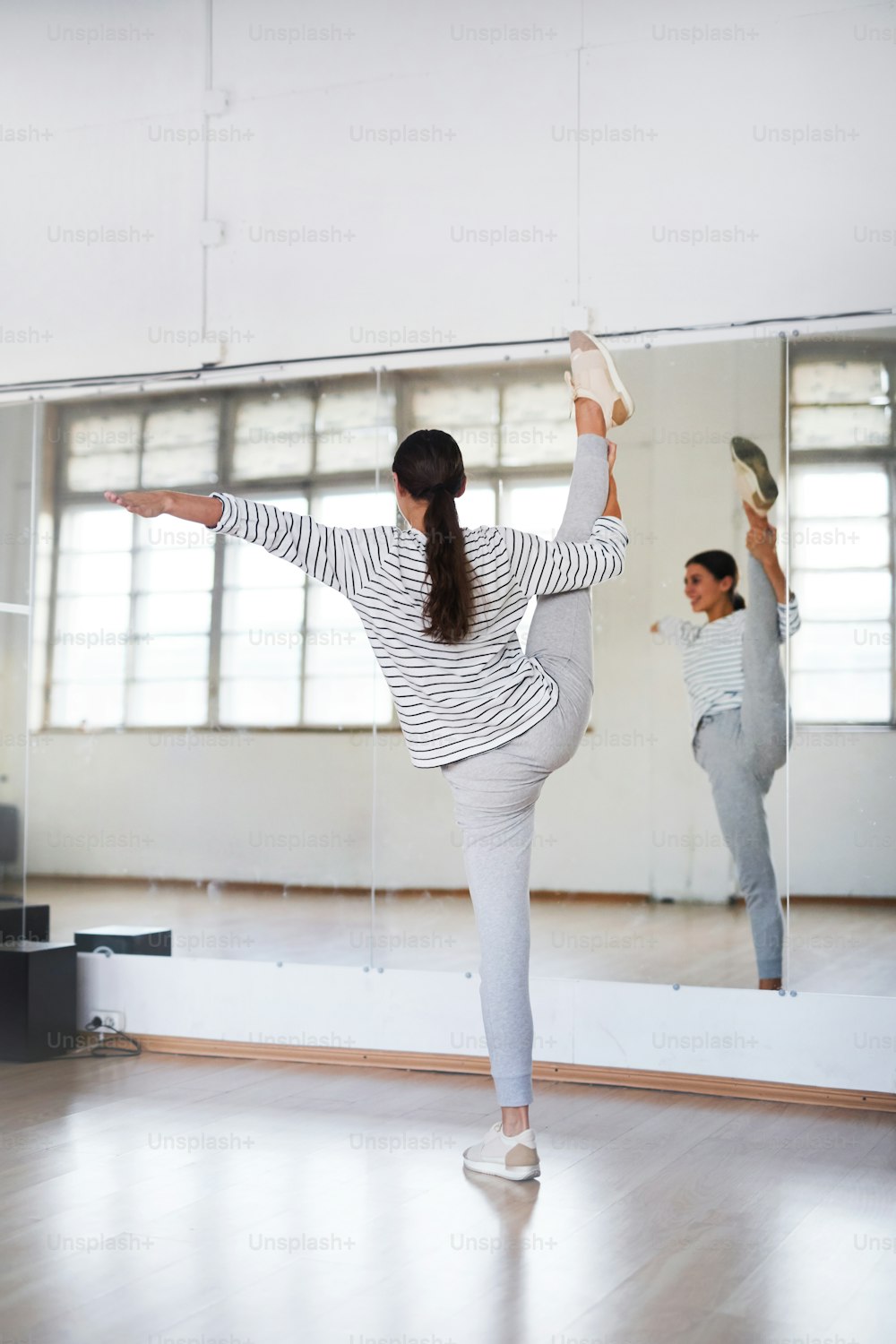 Joven bailarina haciendo ejercicio de estiramiento para brazos y piernas frente al espejo del estudio durante la clase