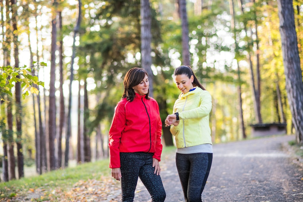 Two female runners with smartwatch standing on a road outdoors in forest in autumn nature, measuring or checking the time.
