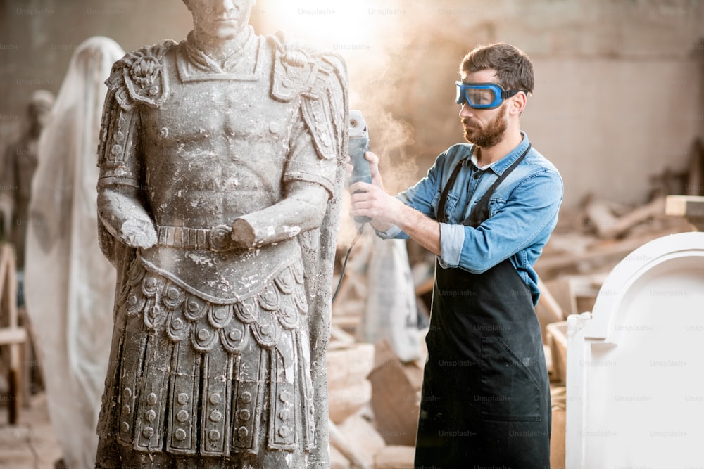 Bildhauer in schützender Arbeitsware schleifende Steinskulptur mit Elektroschleifer im alten Atelier mit Staub