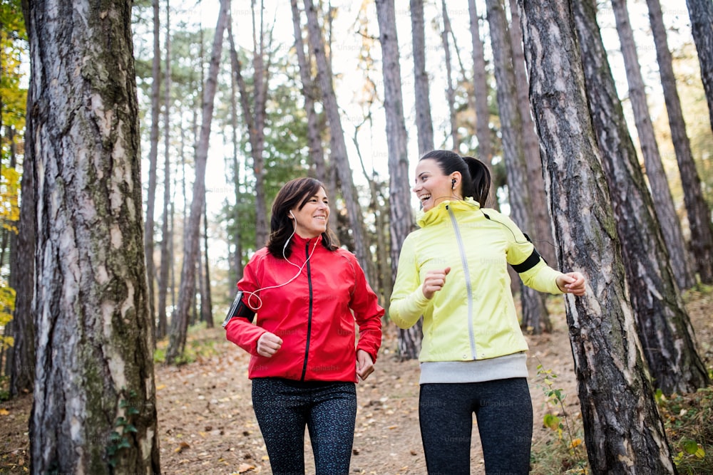 Zwei Läuferinnen mit Kopfhörern und Smartphones im Armband joggen draußen im Wald in der Herbstnatur, im Gespräch.