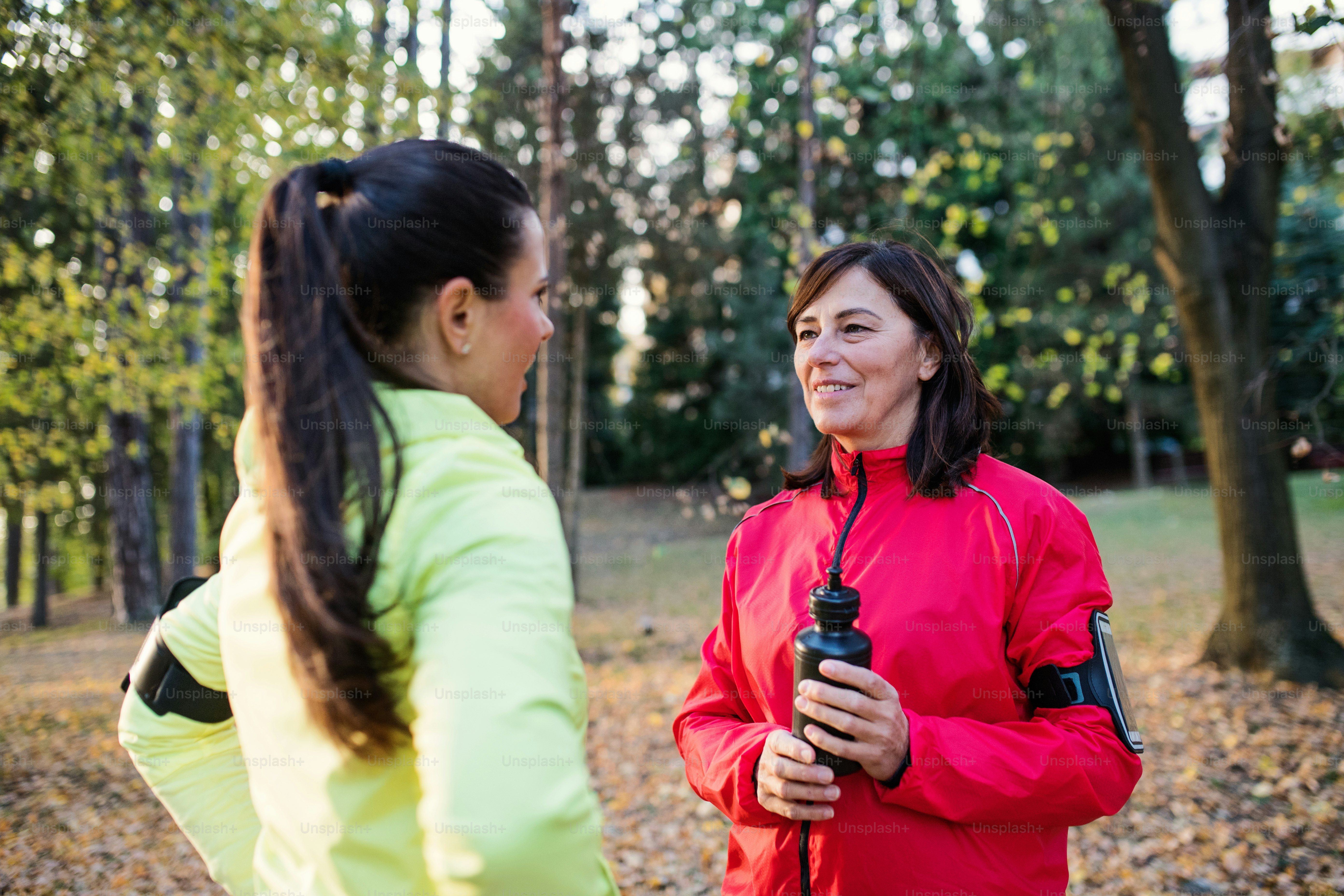 Female runners with water bottle and smartphones standing outdoors in forest, talking when resting.