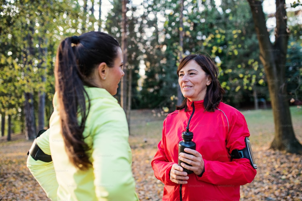 Female runners with water bottle and smartphones standing outdoors in forest, talking when resting.