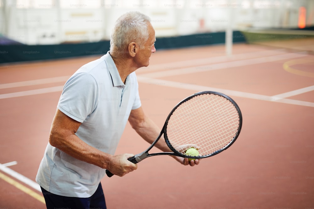 Jugador de tenis envejecido en ropa deportiva sosteniendo la raqueta y la pelota mientras apunta a su opuesto durante el juego