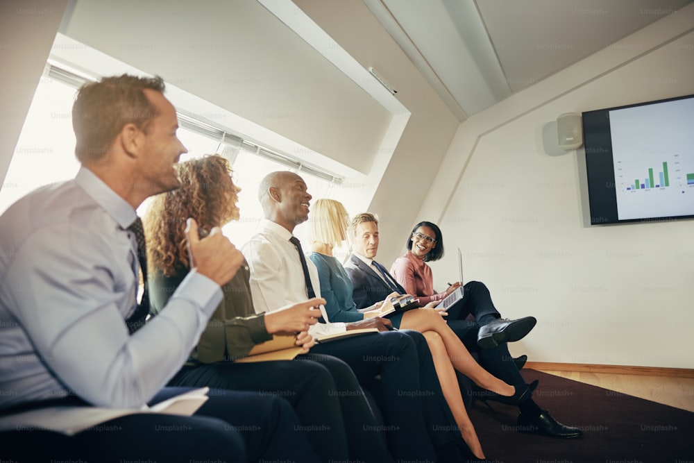 Diverse group of smiling businesspeople sitting on a sofa in an office watching a presentation on a monitor