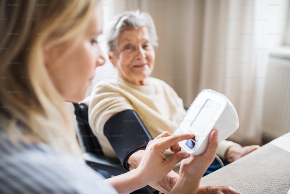 Un giovane visitatore della salute che misura la pressione sanguigna di una donna anziana in sedia a rotelle a casa.