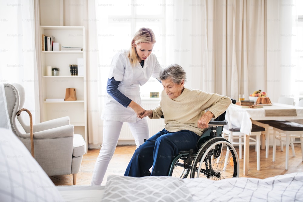 노인 여성이 집에서 휠체어에서 일어설 수 있도록 돕는 젊은 건강 방문자.