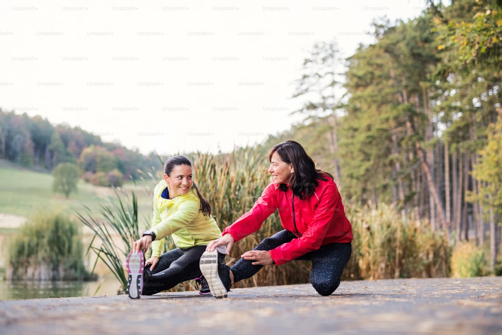 두 명의 활동적인 여성 주자가 달리기 후 가을 자연 속에서 공원에서 야외에서 다리를 뻗고 있습니다.
