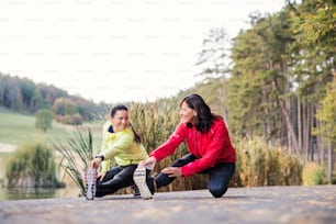 Two active female runners stretching legs outdoors in park in autumn nature after the run.