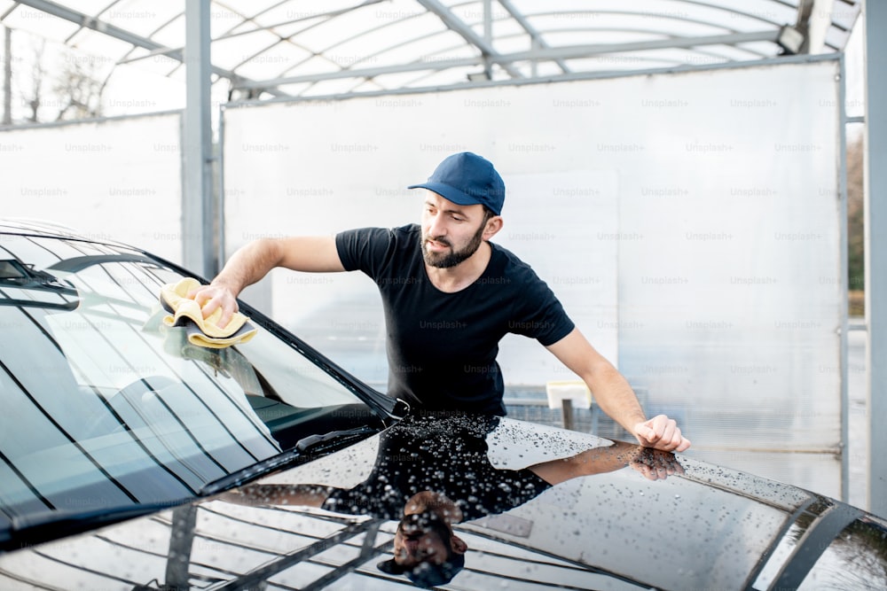 Professional washer in t-shirt and cap wiping windshield with yellow microfiber at the open air car wash