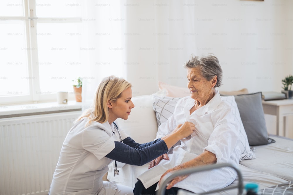 Ein junger Gesundheitsbesucher hilft einer glücklichen, kranken älteren Frau, die zu Hause auf dem Bett sitzt.