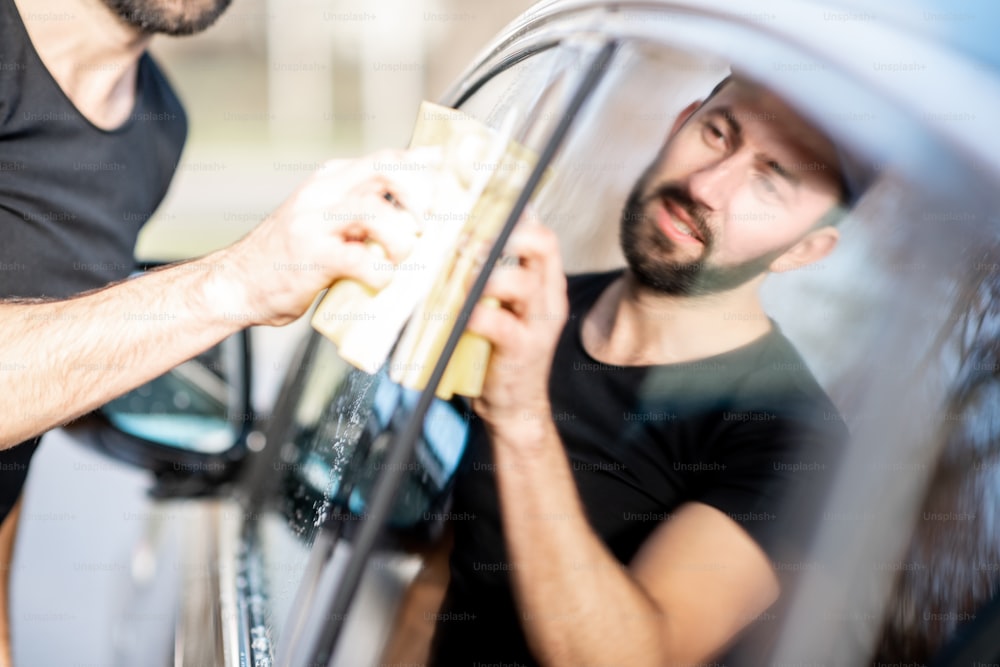 Professionelle Waschmaschine wischt ein Autofenster mit gelbem Schwamm ab und wäscht Luxusauto im Freien