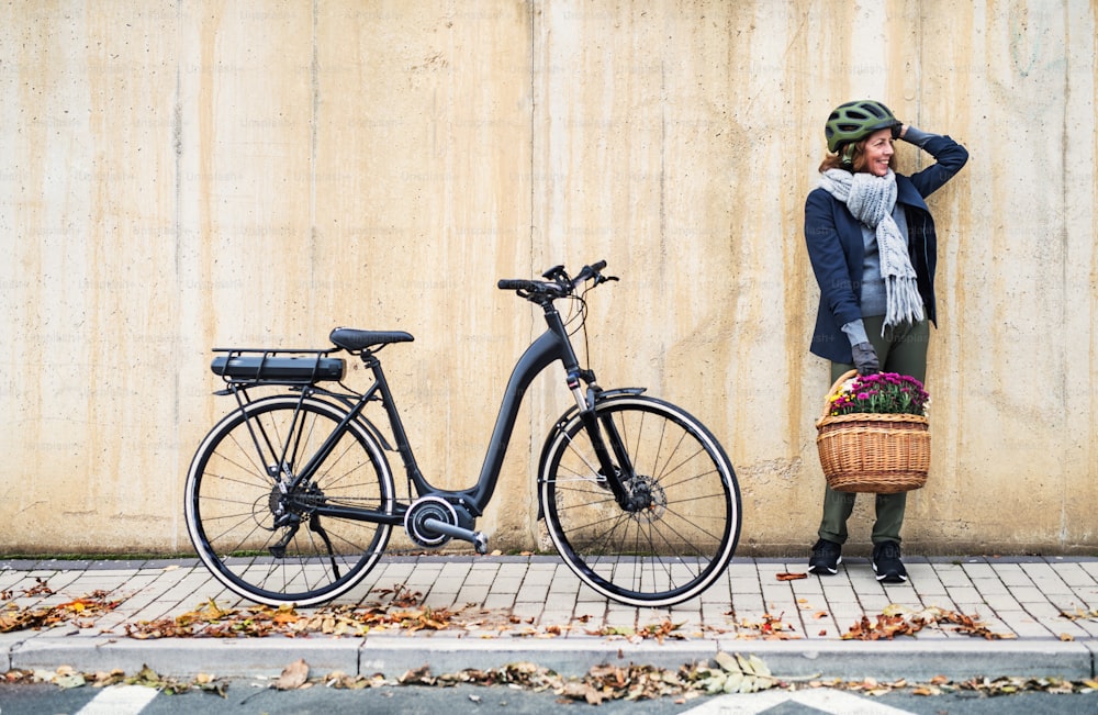 Femme âgée active avec vélo électrique et fleurs dans un panier debout à l’extérieur en ville, appuyée contre un mur en béton. Espace de copie.