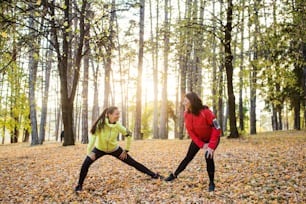 Two active female runners stretching outdoors in forest in autumn nature after the run.