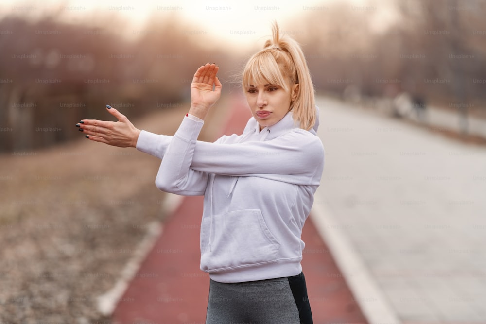 Close up of serious gorgeous blonde Caucasian woman in sportswear and with ponytail stretching arms before running.