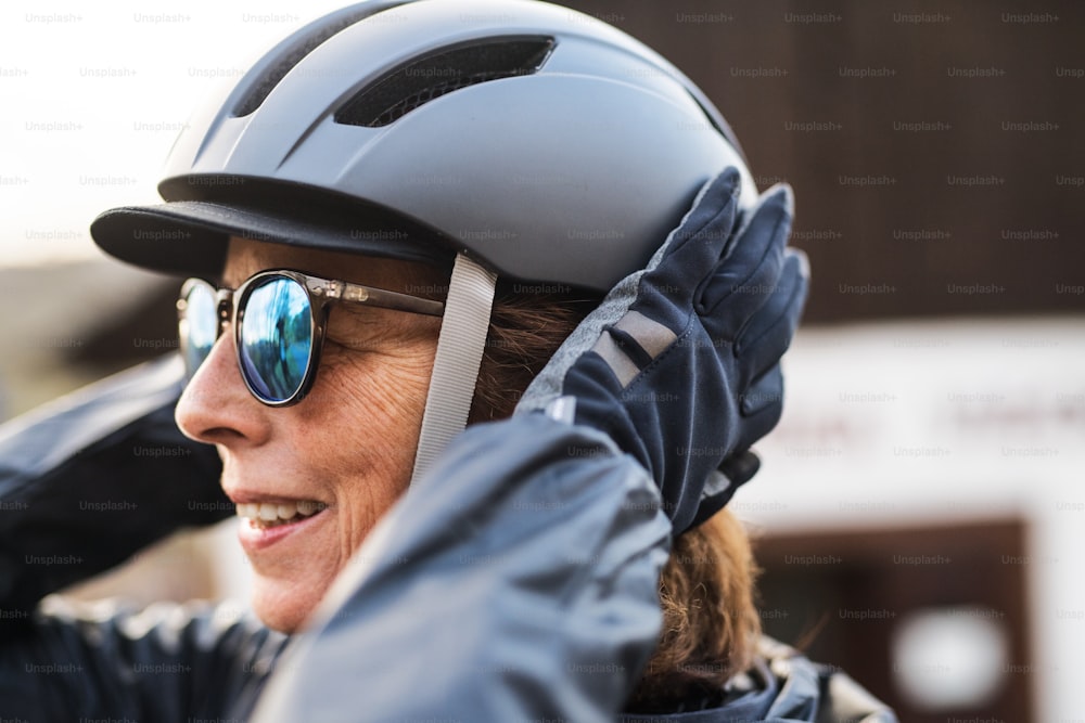 A close-up of active senior woman standing outdoors, putting on bicycle helmet.