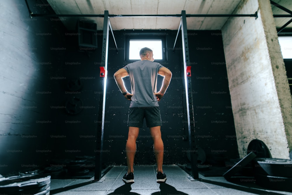 Muskulöser Mann in Sportkleidung, der sich auf Klimmzüge vorbereitet, während er mit den Händen in den Hüften im Fitnessstudio steht. Zurück gedreht.