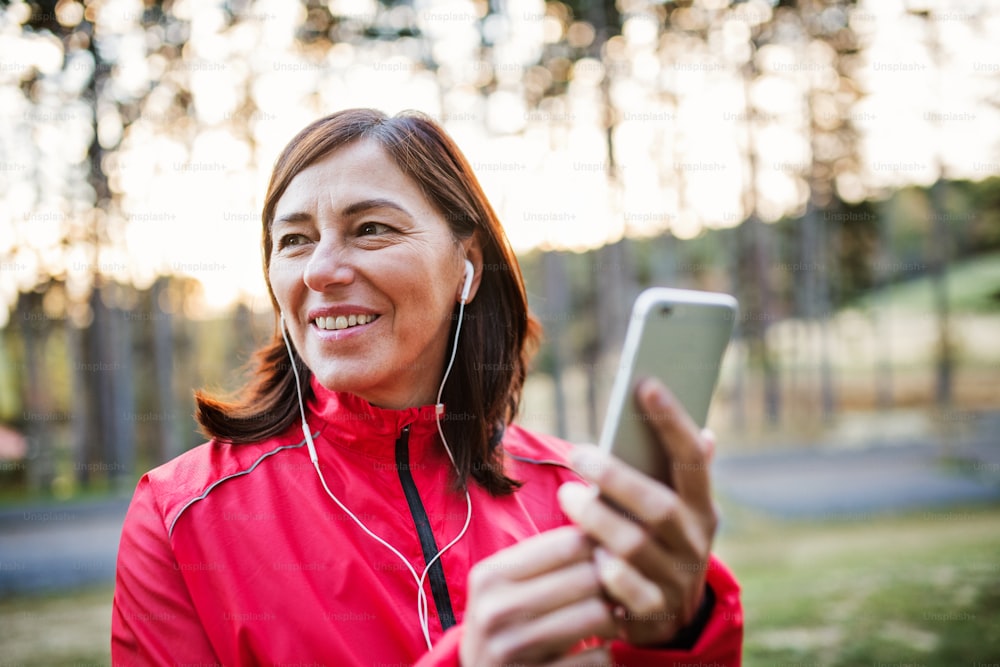 秋の自然の中、屋外でイヤホンとスマートフォンを持ち、音楽を聴きながら走る高齢の女性ランナー。