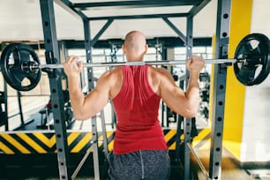 Young bald caucasian bodybuilder lifting barbells while standing in the gym. Backs turned.