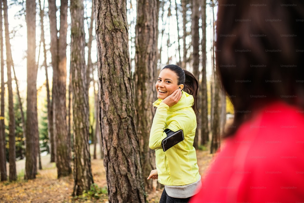 Deux coureuses avec des écouteurs et des smartphones dans le brassard font du jogging en plein air dans la forêt dans la nature d’automne, en train de parler.