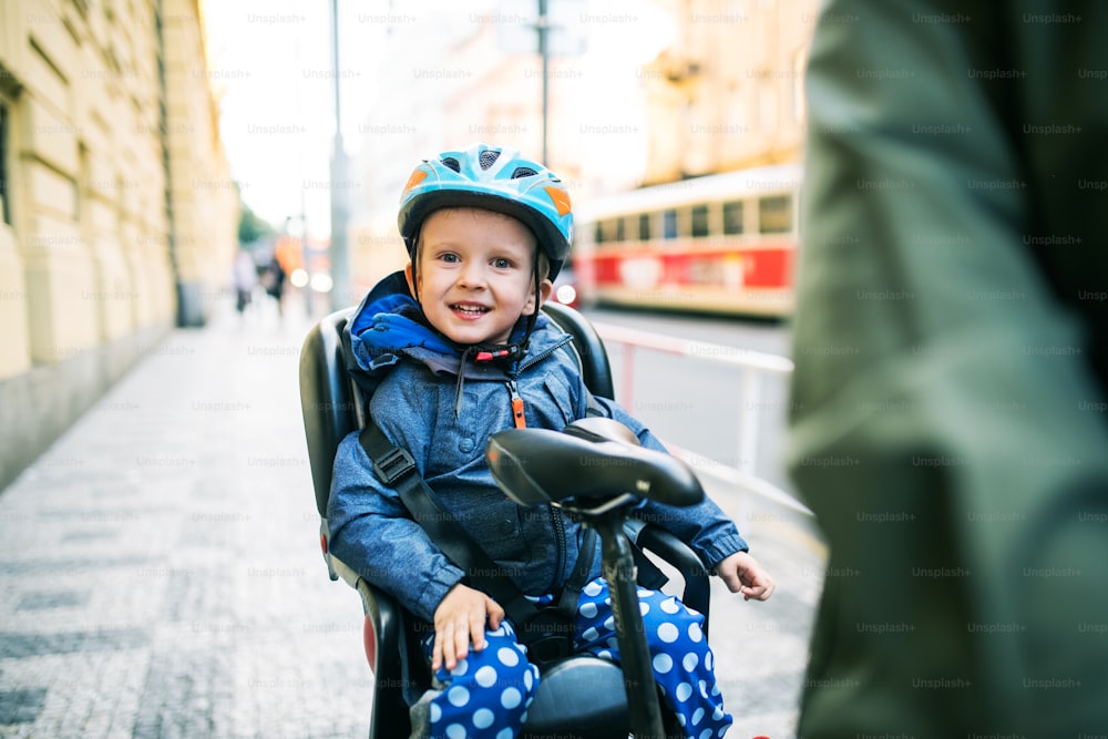 Un niño pequeño con casco sentado en el asiento de la bicicleta con un padre irreconocible al aire libre en una calle de la ciudad.