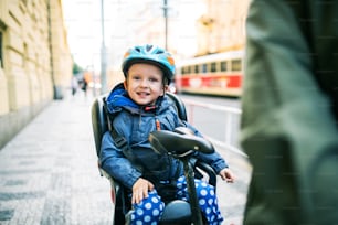 Un petit garçon en bas âge avec un casque assis dans un siège de vélo avec son père méconnaissable à l’extérieur dans une rue de la ville.