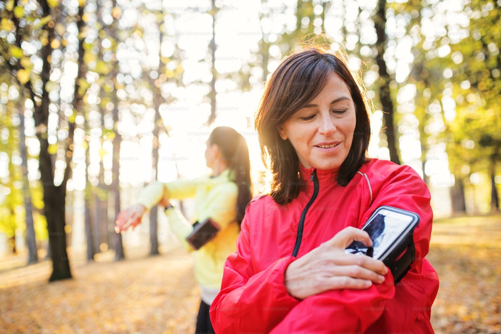Two female runners with smartphone standing outdoors in forest in autumn nature, measuring time.