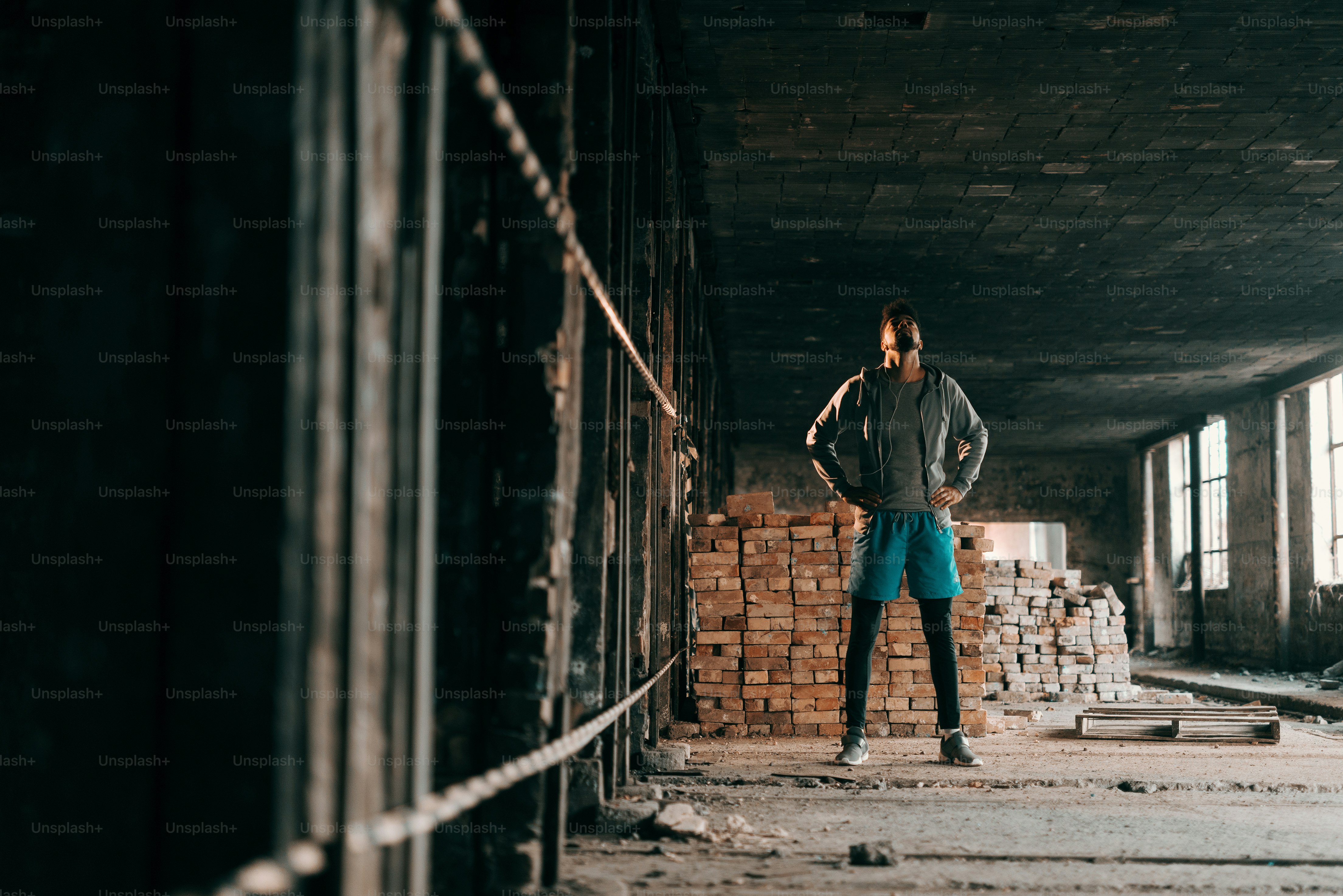 Afroamerikanischer Mann, der aufblickt und Händchen in den Hüften hält, während er in einer alten Ziegelei steht. Urbanes Lifestyle-Konzept.