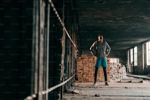 Hombre afroamericano mirando hacia arriba y tomándose de la mano en las caderas mientras está de pie en una vieja fábrica de ladrillos. Concepto de estilo de vida urbano.