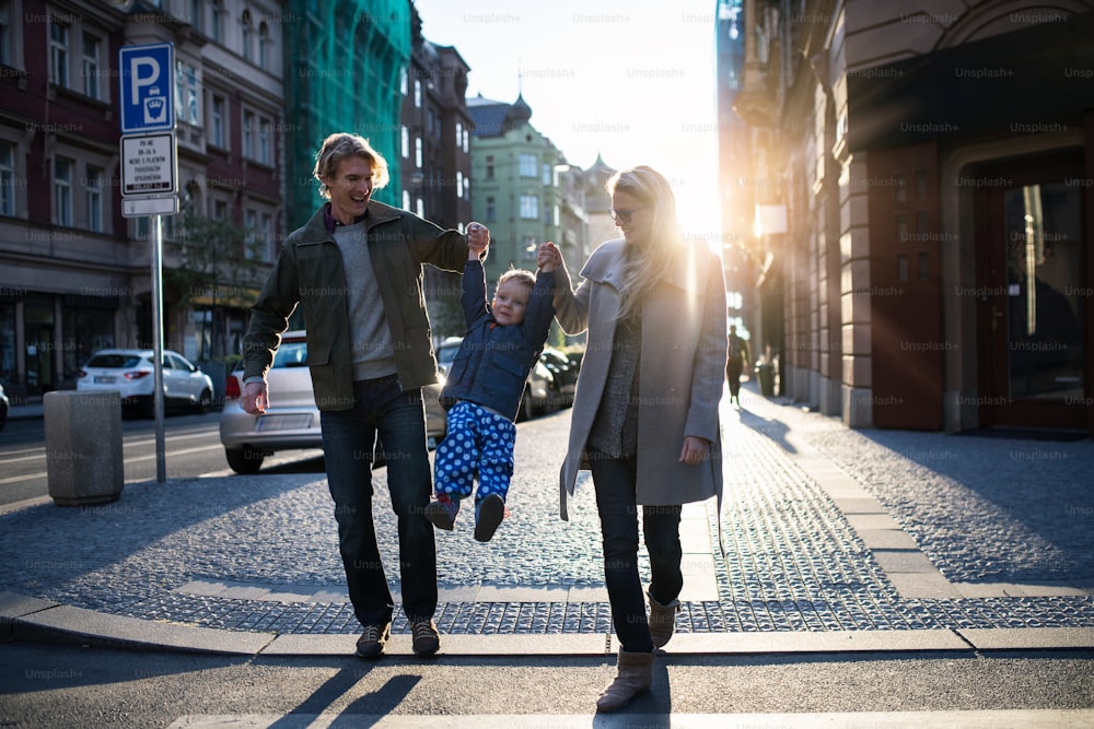 Un petit garçon en bas âge avec ses parents traversant une route à l’extérieur en ville au coucher du soleil, se tenant la main.